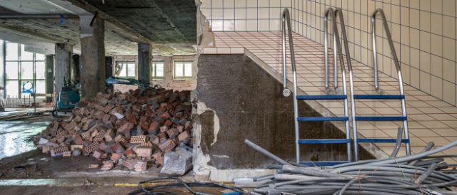 Baustelle Schwimmbad Rialto: rechts im Bild ist ein Wandbild mit gefliester Abbildung zusehen. Davor stehen zwei demontierte Badeleitern. Links im Bild sind Trümmer, Rohre und Baumaterialien zu sehen. Im Hintergrund ein Minibagger und Fenster.